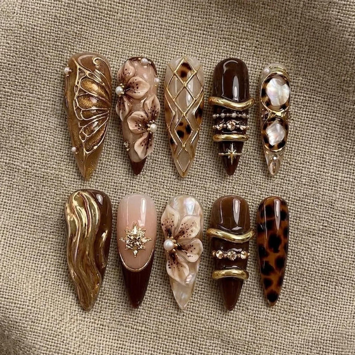 A set of ten almond-shaped press-on nails featuring brown, gold, and cream tones. Designs include 3D floral accents, gold butterfly wings, leopard print patterns, metallic gold swirls, pearl embellishments, and rhinestone details.