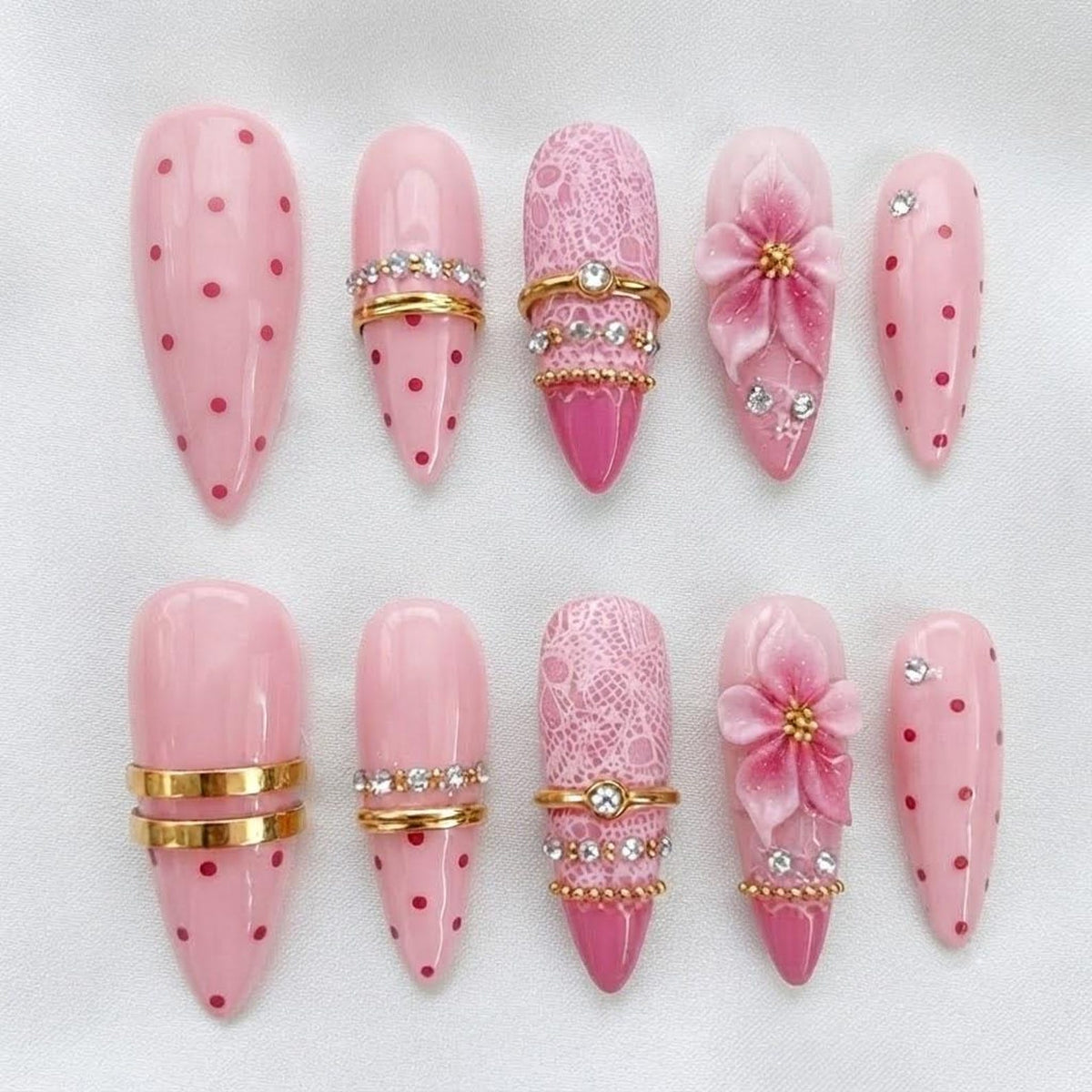 A set of ten press-on nails in varying shades of pink, featuring designs such as red polka dots, gold-toned metal bands, white lace patterns, and three-dimensional pink floral accents with small embedded clear rhinestones.