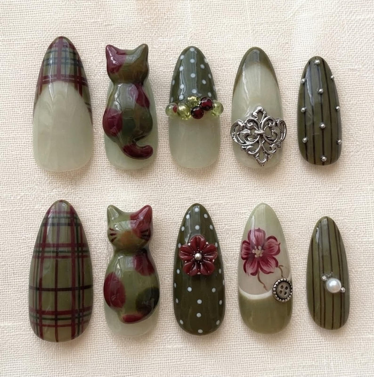 A set of ten almond-shaped press-on nails featuring olive green, cream, and maroon tones. Designs include plaid patterns, 3D cat figures, white polka dots with beads or flowers, silver filigree, painted floral art, buttons, and pearls.