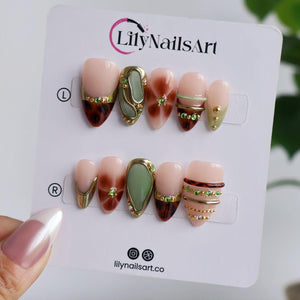 A hand holds a white display card with ten almond press-on nails set in two rows, the top row marked L and the bottom marked R. Designs include tortoiseshell tips, a sage green cabochon, gold rims, and green gems on nude bases.