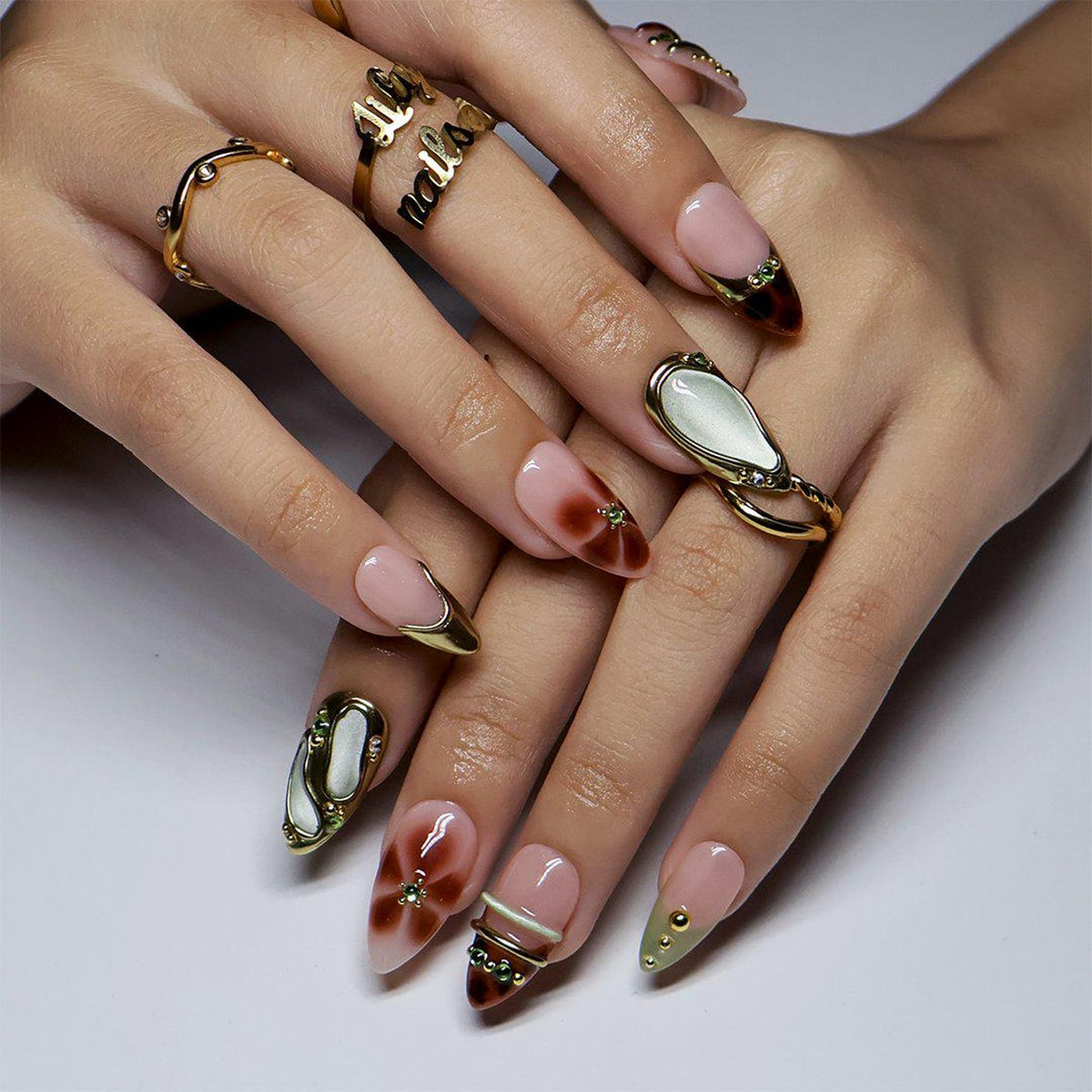 Close-up of two hands resting one over the other, nails angled toward the camera. Sheer nude almond nails with tortoiseshell French tips, raised sculpted gold borders, and a pale jade cabochon panel sitting above the nail surface.
