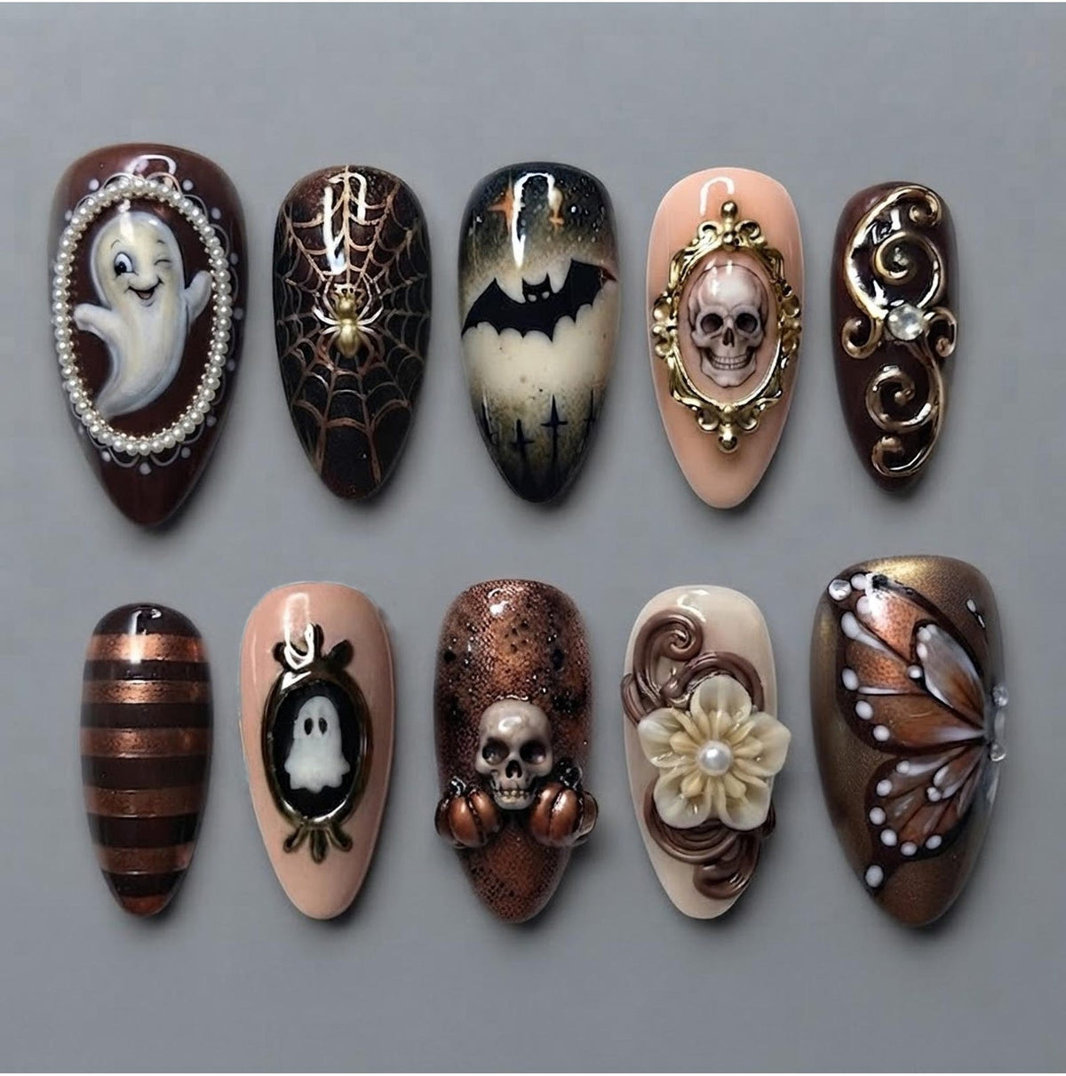 A set of ten almond-shaped press-on nails featuring Halloween designs including a ghost in a pearl frame, a spider web, a bat, a skull in a gold frame, metallic swirls, brown stripes, a ghost in a frame, a skull with pumpkins, a flower, and a butterf