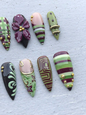 A set of eight almond-shaped press-on nails featuring various designs in shades of olive green, deep plum, and cream. The nails are decorated with hand-painted plaid patterns, floral motifs, geometric lines, and small gold metallic studs.