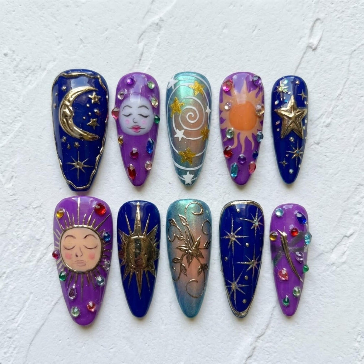 A set of ten almond-shaped press-on nails featuring celestial designs. The nails are decorated with gold metallic moons, suns, and stars, painted faces, swirls, and colorful rhinestones on deep blue, purple, and iridescent backgrounds.