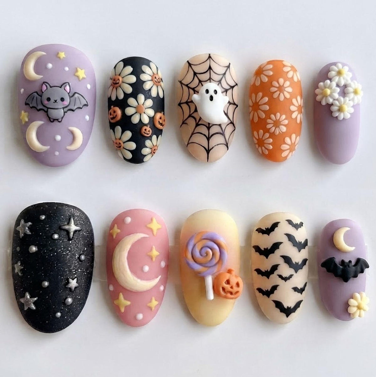 A set of ten press-on nails featuring Halloween-themed designs including a bat, crescent moons, stars, daisies, pumpkins, a spider web, a ghost, a lollipop, and flying bats in a color palette of lavender, black, orange, and cream.
