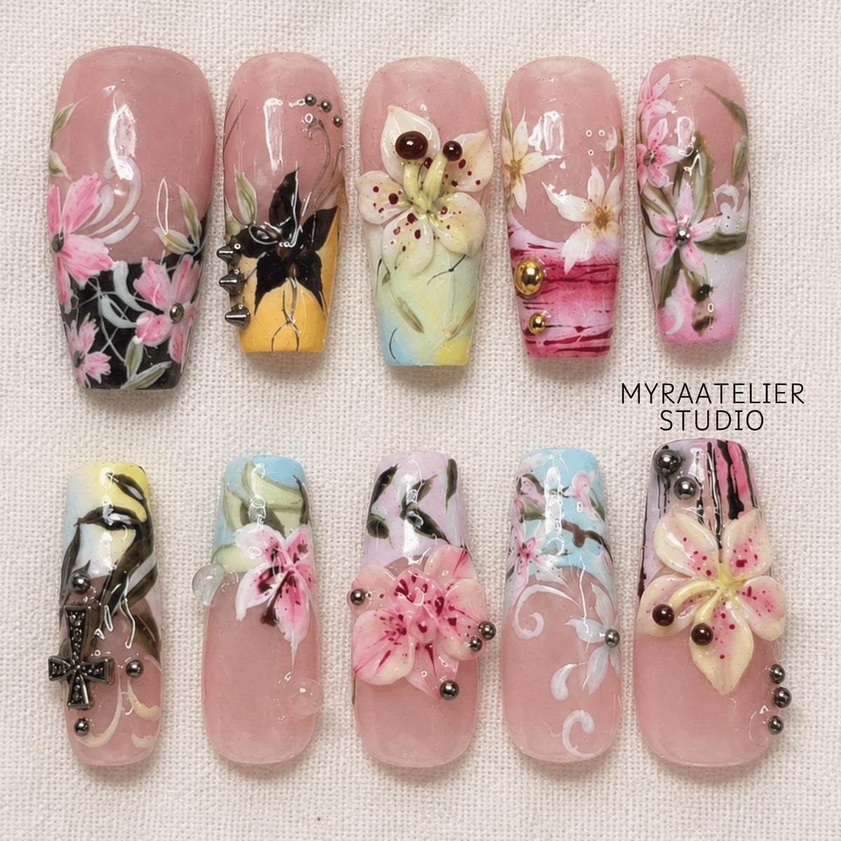 A set of ten press-on nails featuring a nude base with intricate hand-painted floral designs, 3D flower embellishments, metallic studs, and black cross accents. The text MYRAATELIER STUDIO is printed in black on the right side.