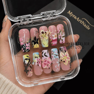 A set of ten press-on nails in a clear plastic case, featuring a nude base with intricate hand-painted floral designs, 3D flower accents, metallic studs, and a small cross charm. The text MyraArtNails and MYRAATELIER STUDIO is visible.