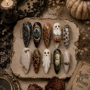A set of ten almond-shaped press-on nails featuring Halloween-themed designs including black lace, leopard print, gold skull accents, white ghosts, a pumpkin, and a framed ghost illustration, all arranged on a textured display tray.