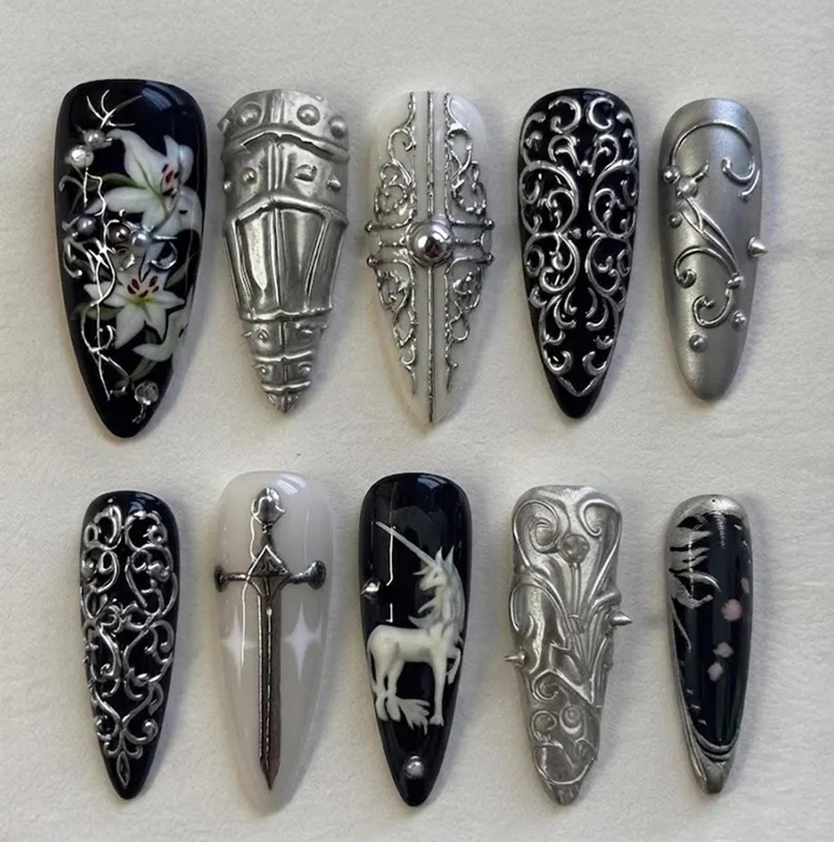 A set of ten almond-shaped press-on nails featuring intricate designs in black, white, and metallic silver. The collection includes motifs such as white lilies, silver armor plating, ornate filigree, a silver sword, and a white unicorn.