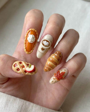 Five fingernails featuring 3D nail art designs. One nail shows an orange pumpkin pie slice with whipped cream, another a coffee cup with latte art, a third a golden croissant, a fourth an apple pie slice, and the last a maple leaf.