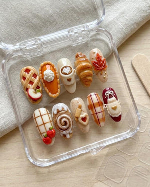 A set of ten press-on nails featuring 3D autumn-themed designs including a lattice apple pie, pumpkin pie, latte art, croissant, maple leaf, plaid patterns, a cinnamon roll, a rolling pin with a heart cookie, and a kitchen apron.