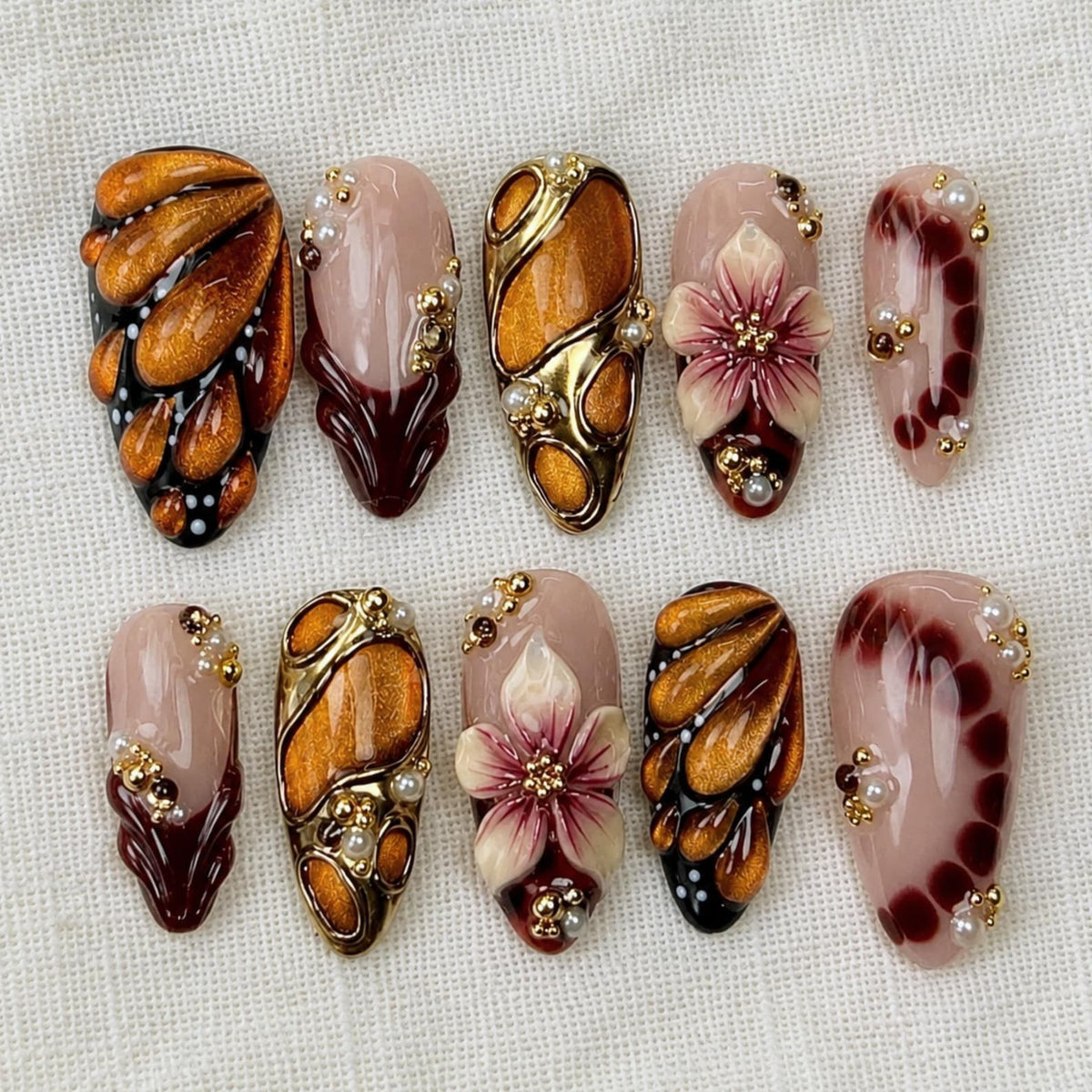 A set of ten press-on nails featuring a mix of designs including metallic gold and copper teardrop patterns, deep burgundy accents, and three-dimensional floral motifs, all embellished with small gold and white spherical beads.