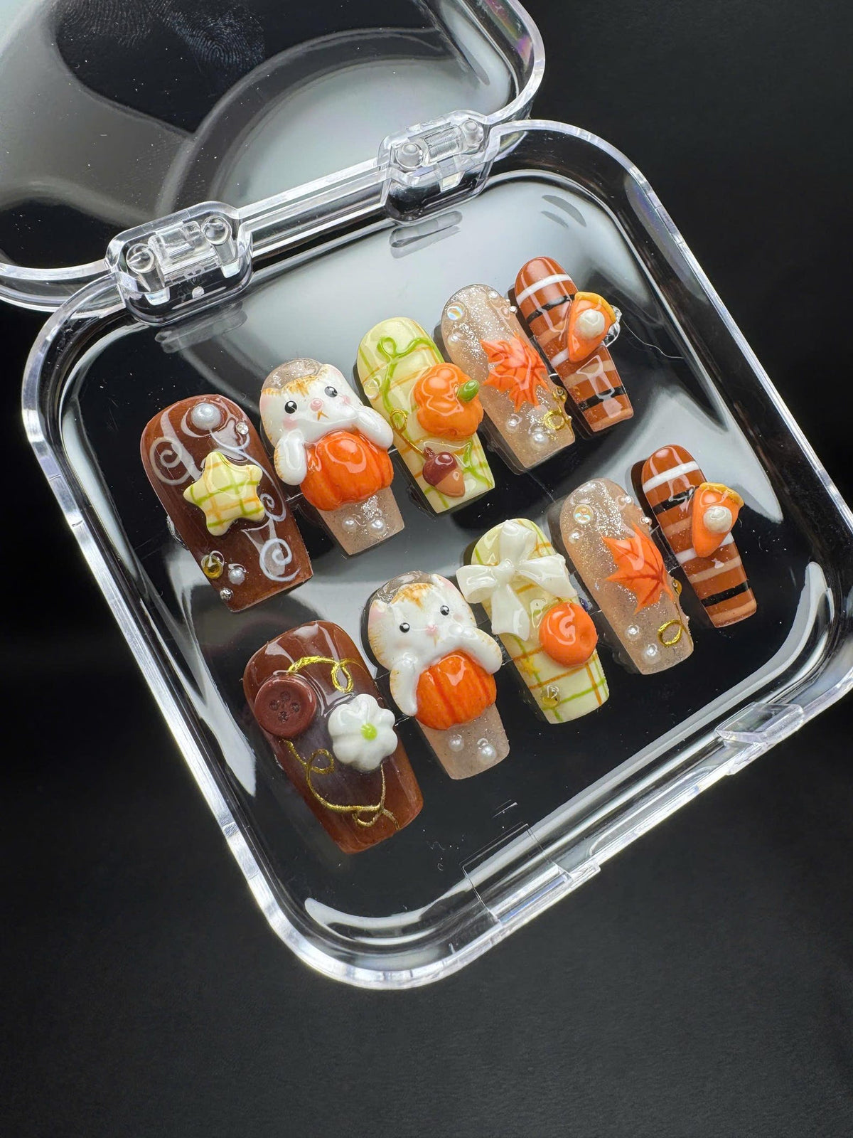 A set of ten press-on nails in a clear plastic case, featuring autumn-themed designs including 3D pumpkins, white rabbits, maple leaves, plaid patterns, bows, buttons, and glitter accents in shades of orange, brown, white, and gold.