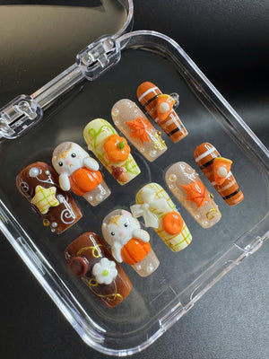 A set of ten press-on nails featuring autumn-themed designs including orange pumpkins, white rabbits, maple leaves, plaid patterns, and small pie slices. The nails are decorated with 3D charms, glitter, and pearls in a clear plastic case.