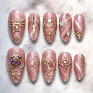 A set of ten almond-shaped press-on nails in a shimmering rose gold finish. The designs feature gold-toned 3D emblems including the Triforce, a sword, a shield with a red bird, a fairy, a profile of a character, and an eye symbol.