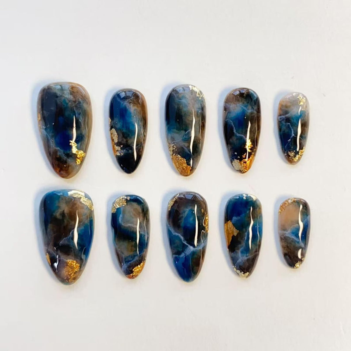 A set of ten press-on nails featuring a glossy finish with a marbled pattern in shades of deep blue, black, and brown, accented with small flecks of gold leaf throughout the design. The nails are arranged in two rows of five.