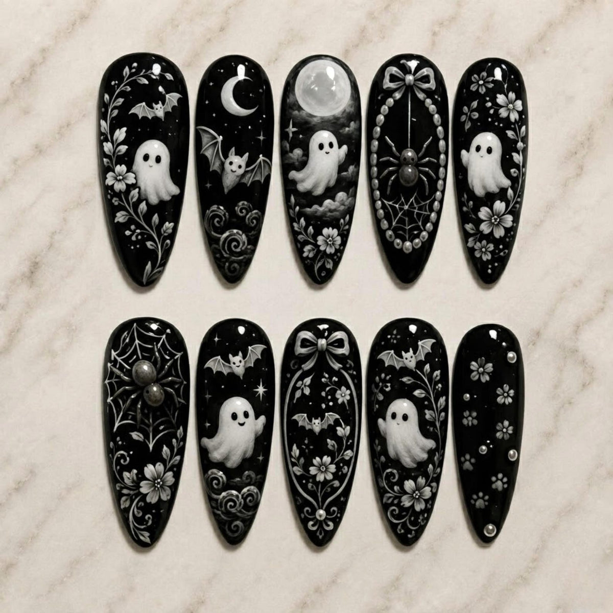 A set of ten almond-shaped press-on nails with a glossy black base. The designs feature white hand-painted motifs including ghosts, bats, spiders, spiderwebs, crescent moons, full moons, bows, floral vines, and small circular accents.