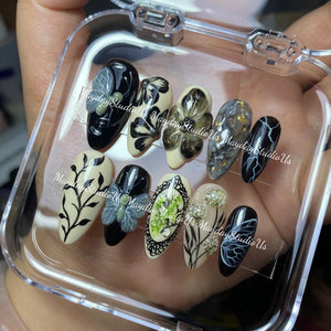 A set of ten almond-shaped press-on nails in a clear plastic case. The designs include black and cream bases with hand-painted botanical motifs, 3D butterfly accents, marble patterns with gold flakes, lightning bolts, and lace details.
