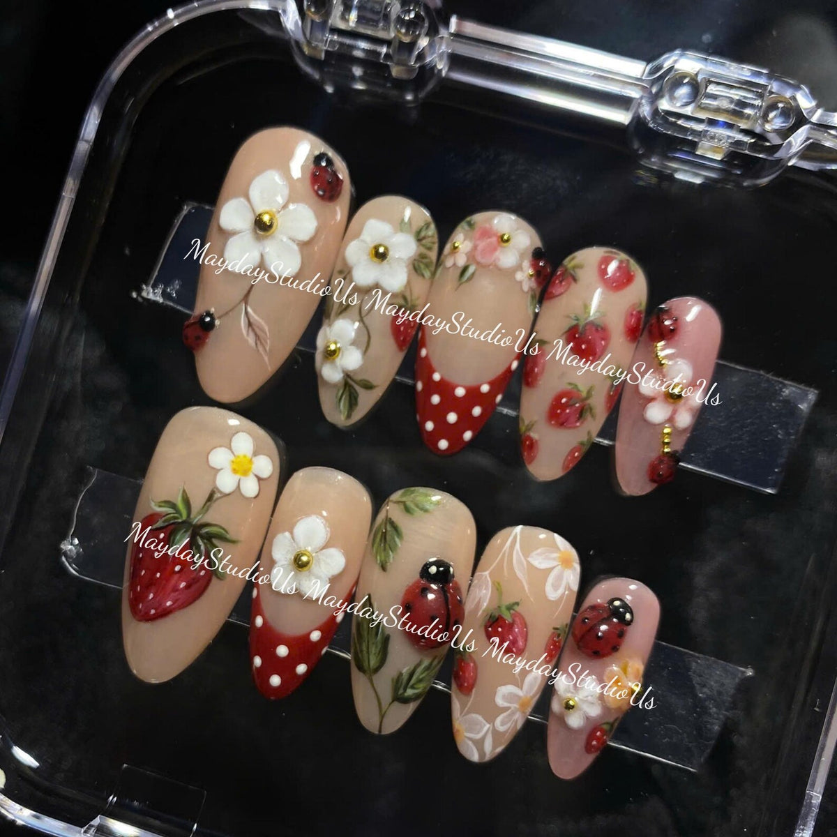 A set of ten almond-shaped press-on nails featuring a nude base with hand-painted designs including red strawberries, white flowers with gold centers, green leaves, and red ladybugs with black spots, plus red tips with white polka dots.