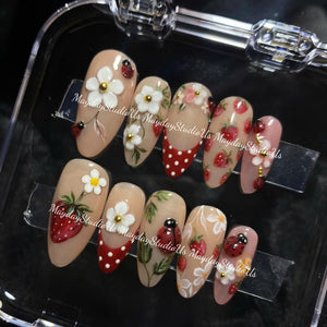 A set of ten almond-shaped press-on nails featuring a nude base with hand-painted designs including red strawberries, white flowers with gold centers, green leaves, and red ladybugs with black spots, plus red tips with white polka dots.