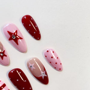 A set of almond-shaped press-on nails featuring a mix of designs. Some nails are deep red with white polka dots, while others are light pink with red star accents and small clear rhinestones. One nail has a French tip style.