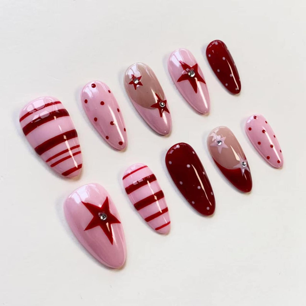A set of ten almond-shaped press-on nails featuring a variety of designs in shades of red and light pink. The patterns include horizontal stripes, polka dots, and red star motifs accented with small, clear, circular rhinestones.