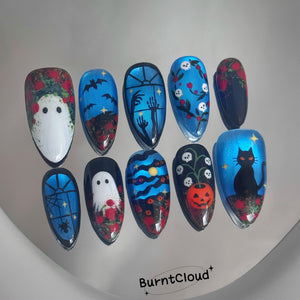 A set of ten almond-shaped press-on nails featuring Halloween-themed artwork including ghosts, bats, skulls, a black cat, a pumpkin, and spiderwebs in shades of blue, black, red, and white. The text BurntCloud is printed at the bottom.