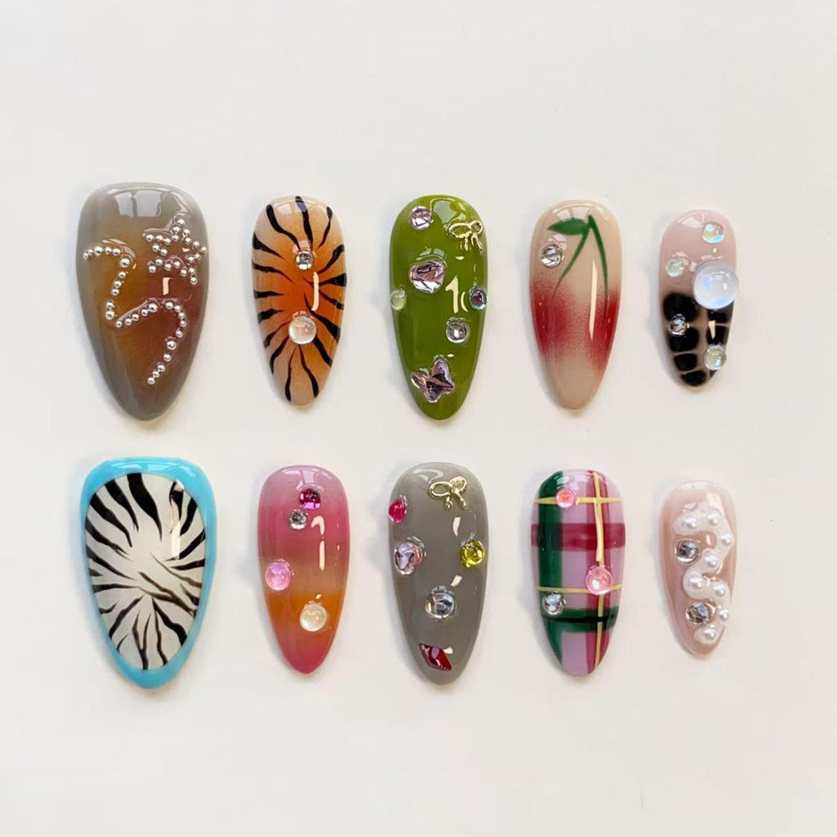 A set of ten press-on nails featuring diverse designs including tiger stripes, plaid patterns, animal prints, and solid colors. The nails are decorated with various embellishments such as small white pearls, clear rhinestones, and gold-toned charms.