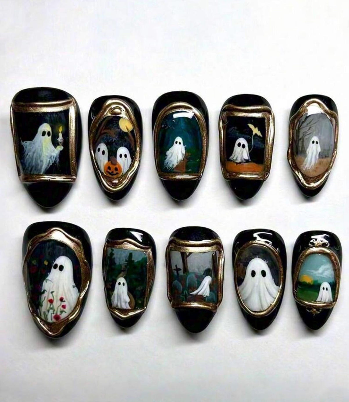 A set of ten press-on nails with a black base and gold-painted frames. Each nail features a unique hand-painted scene of white ghosts in various settings, including a graveyard, a pumpkin patch, and floral fields, with detailed art.