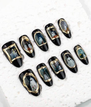 A set of ten press-on nails with a glossy black base. Each nail features a central oval or rectangular frame in metallic gold, enclosing a detailed illustration of a white ghost. The designs vary, showing ghosts in different poses.