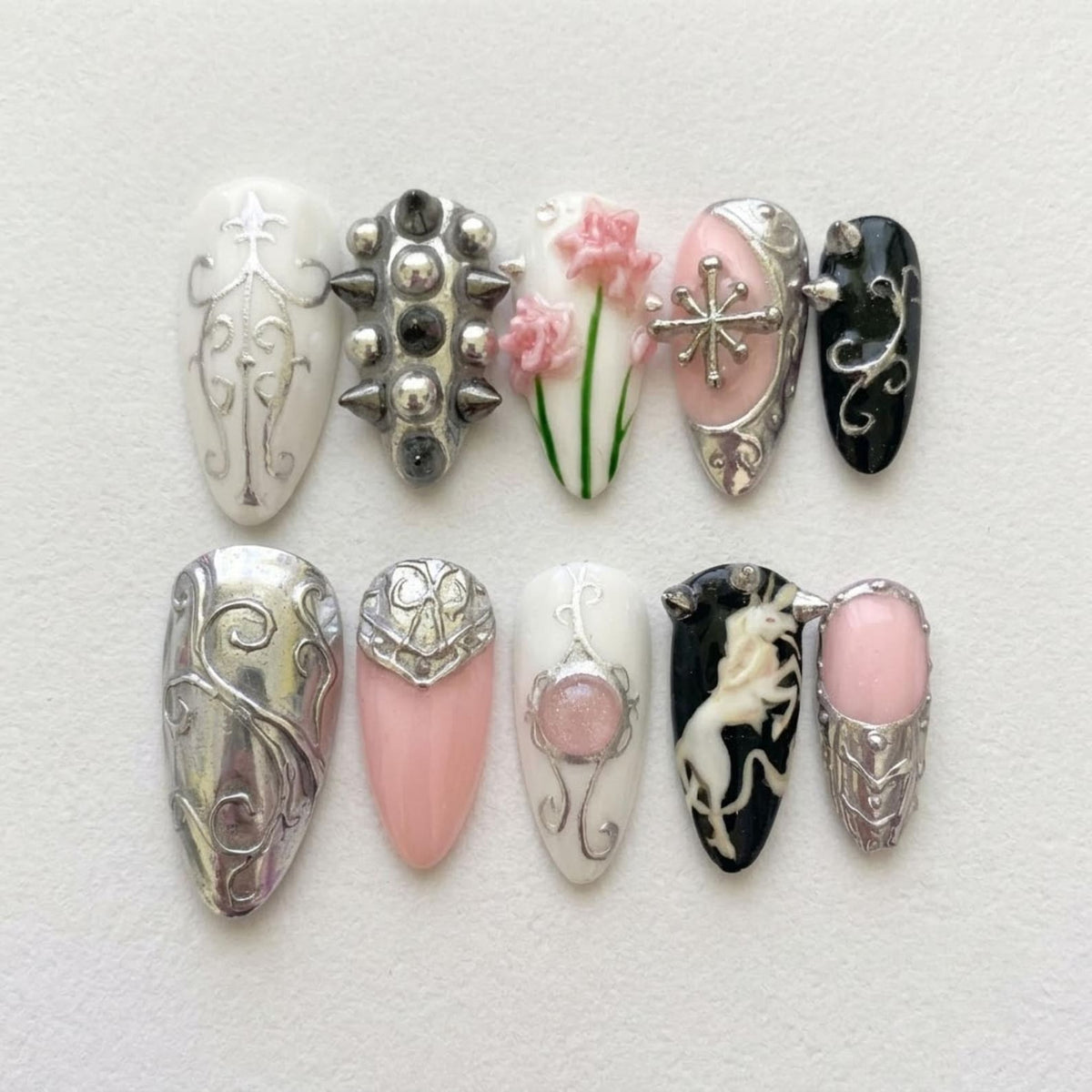 A set of ten press-on nails featuring diverse designs including silver filigree on white, metallic studs, pink floral accents, a silver cross, black with silver swirls, a solid silver finish, pink with silver armor, and a unicorn graphic.