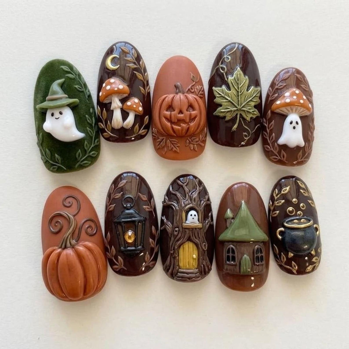 A set of ten oval-shaped press-on nails featuring 3D autumn and Halloween designs. The collection includes motifs of a white ghost in a green hat, mushrooms, a carved pumpkin, a maple leaf, a lantern, a tree house, a cottage, and a cauldron.
