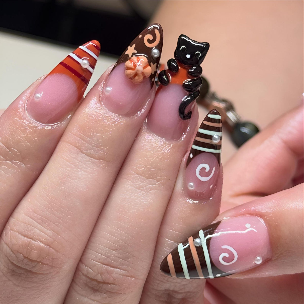 Almond-shaped artificial nails featuring a nude base with brown, orange, and white tips. Designs include horizontal stripes, white swirls, a small 3D orange pumpkin, a black cat figurine, and tiny white pearl accents on each nail.