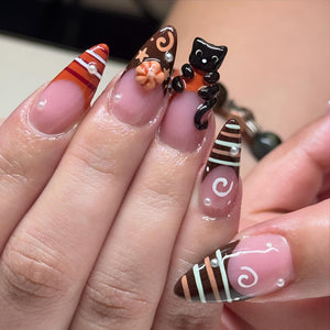 Almond-shaped artificial nails featuring a nude base with brown, orange, and white tips. Designs include horizontal stripes, white swirls, a small 3D orange pumpkin, a black cat figurine, and tiny white pearl accents on each nail.