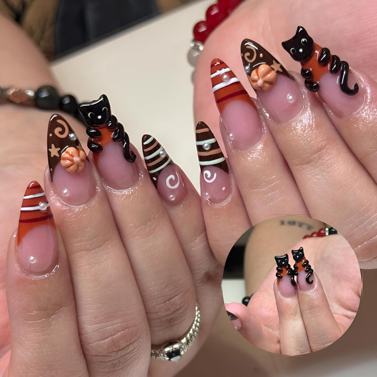 Almond-shaped nails featuring a French manicure style with brown and orange tips. The design includes 3D black cat figures, small orange pumpkins, white swirl accents, and tiny white pearl embellishments on a clear base coat.