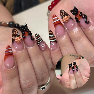 Almond-shaped nails featuring a French manicure style with brown and orange tips. The design includes 3D black cat figures, small orange pumpkins, white swirl accents, and tiny white pearl embellishments on a clear base coat.