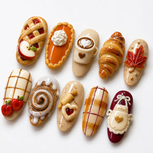 A set of ten oval-shaped nail art charms featuring miniature 3D designs including an apple pie, pumpkin pie with whipped cream, a coffee mug, a croissant, a maple leaf, plaid patterns, cinnamon rolls, a rolling pin, and an apron.
