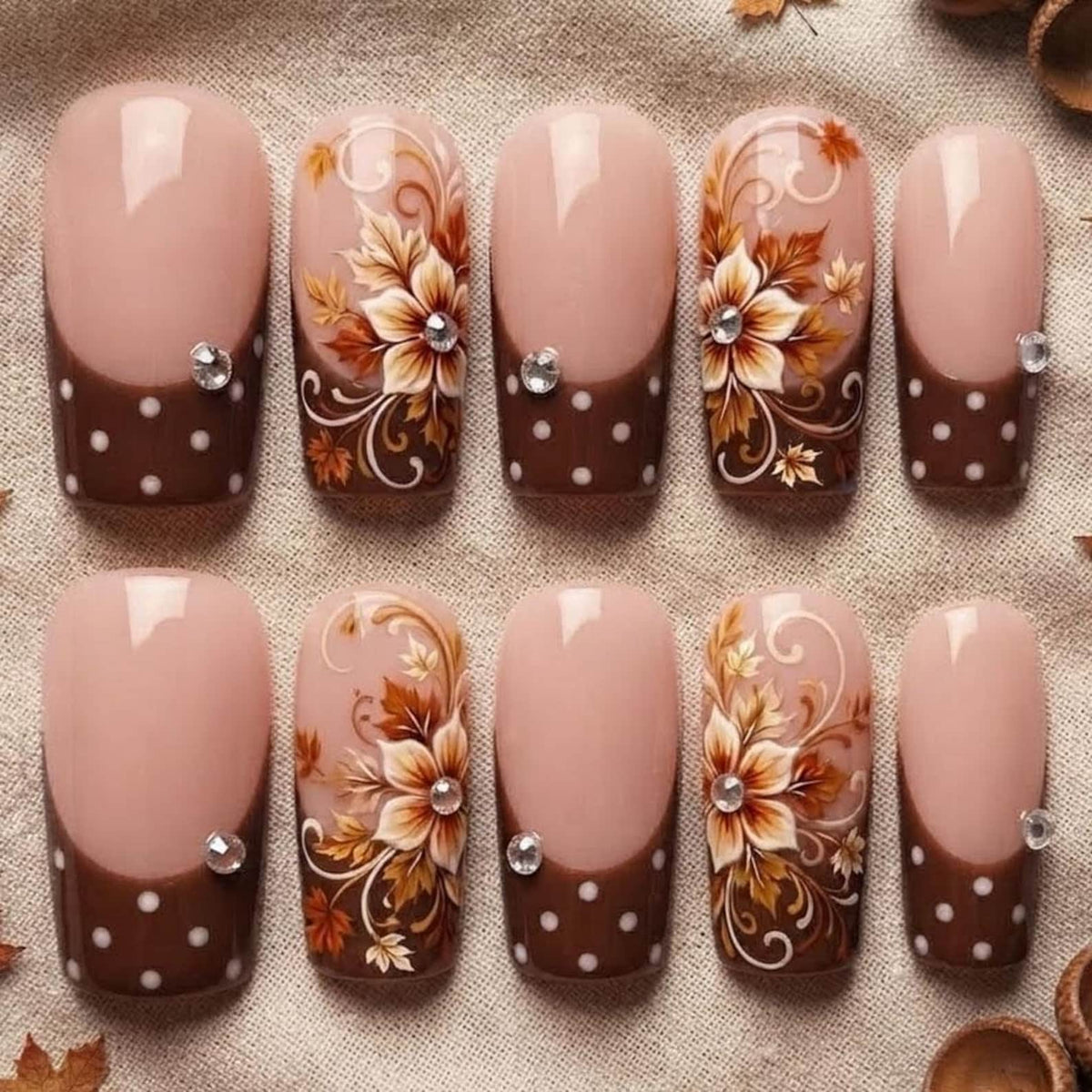 A set of ten press-on nails featuring a nude base with brown tips. Some nails have white polka dots, while others display intricate brown and white floral patterns with swirling leaves. Each nail is accented with a small, clear rhinestone.