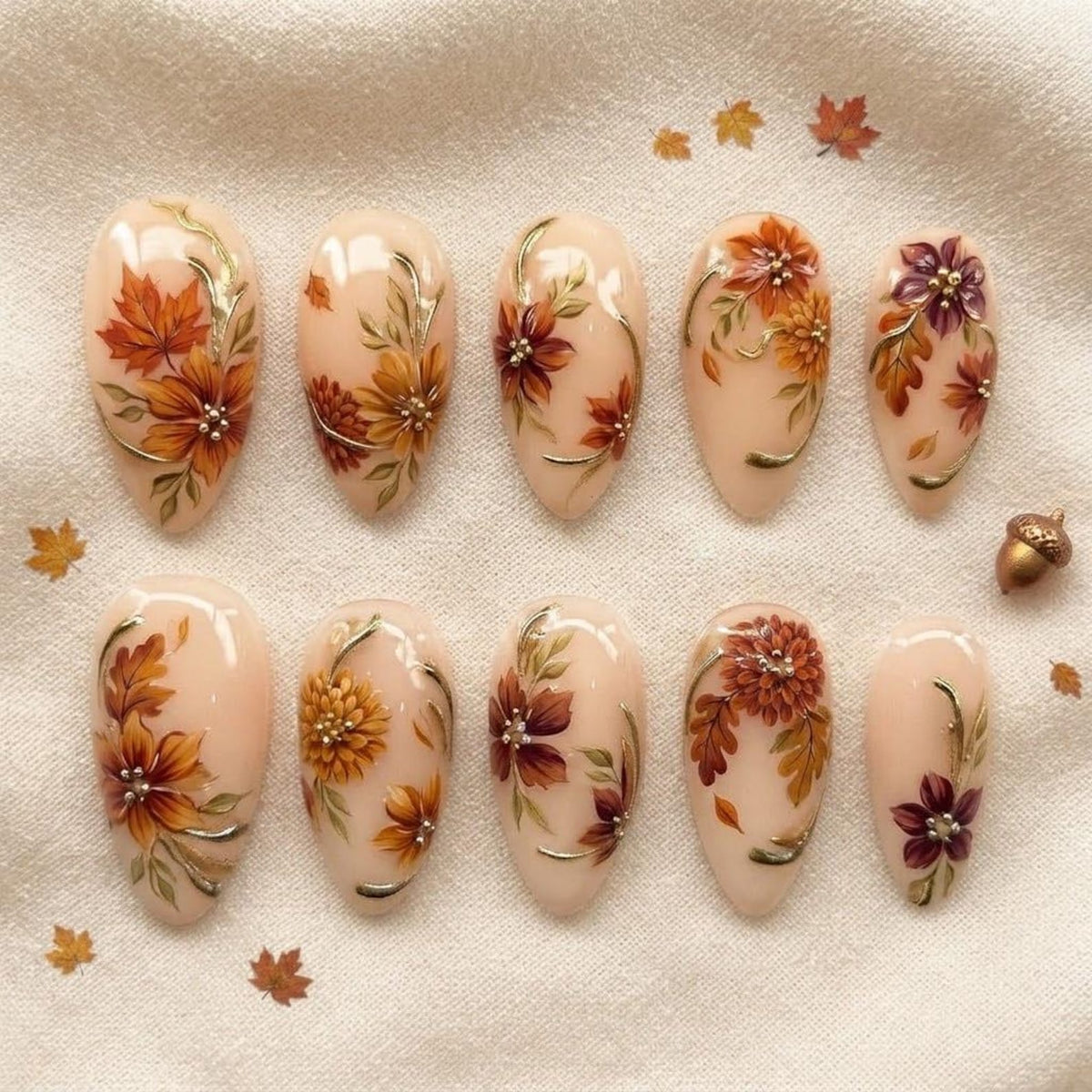 A set of ten almond-shaped press-on nails with a nude base, featuring hand-painted autumn-themed designs including orange, yellow, and purple flowers, maple leaves, and delicate gold metallic accents, arranged in two rows of five.