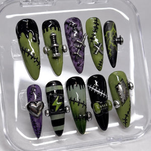 A set of ten press-on nails in a clear plastic case, featuring almond-shaped tips in shades of olive green, black, and purple. Designs include black drip patterns, silver metal studs, lightning bolts, heart charms, and stitched details.
