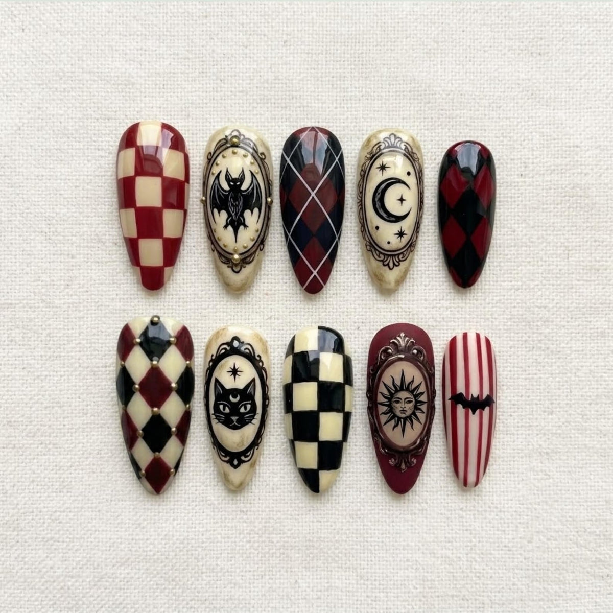 A set of ten almond-shaped press-on nails featuring various designs including red and cream checkerboard, black and red argyle, vertical stripes, and ornate oval frames containing illustrations of a bat, moon, cat, and sun.