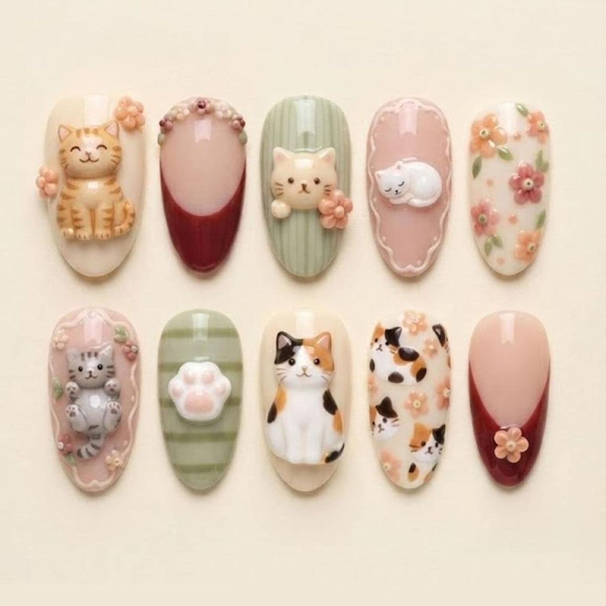 A set of ten almond-shaped press-on nails featuring 3D cat designs, paw prints, and floral accents. The collection includes cream, sage green, and burgundy colors with detailed miniature feline figures and small sculpted flower embellishments.
