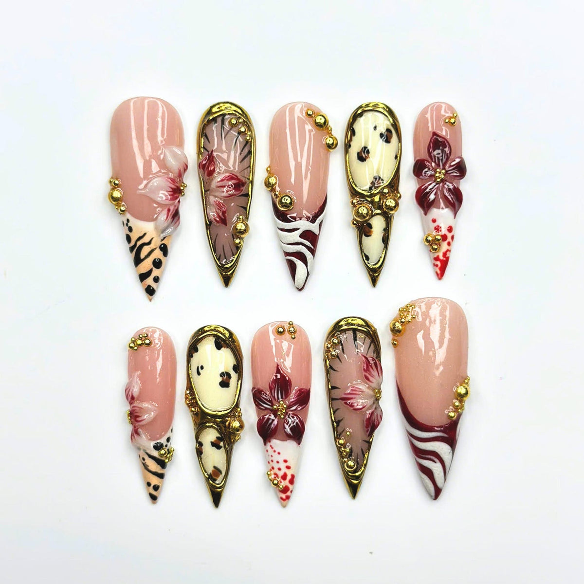 A set of ten stiletto-shaped press-on nails featuring a nude base with intricate designs including hand-painted maroon flowers, black and white animal prints, gold metallic accents, and small gold beads applied to the surface.