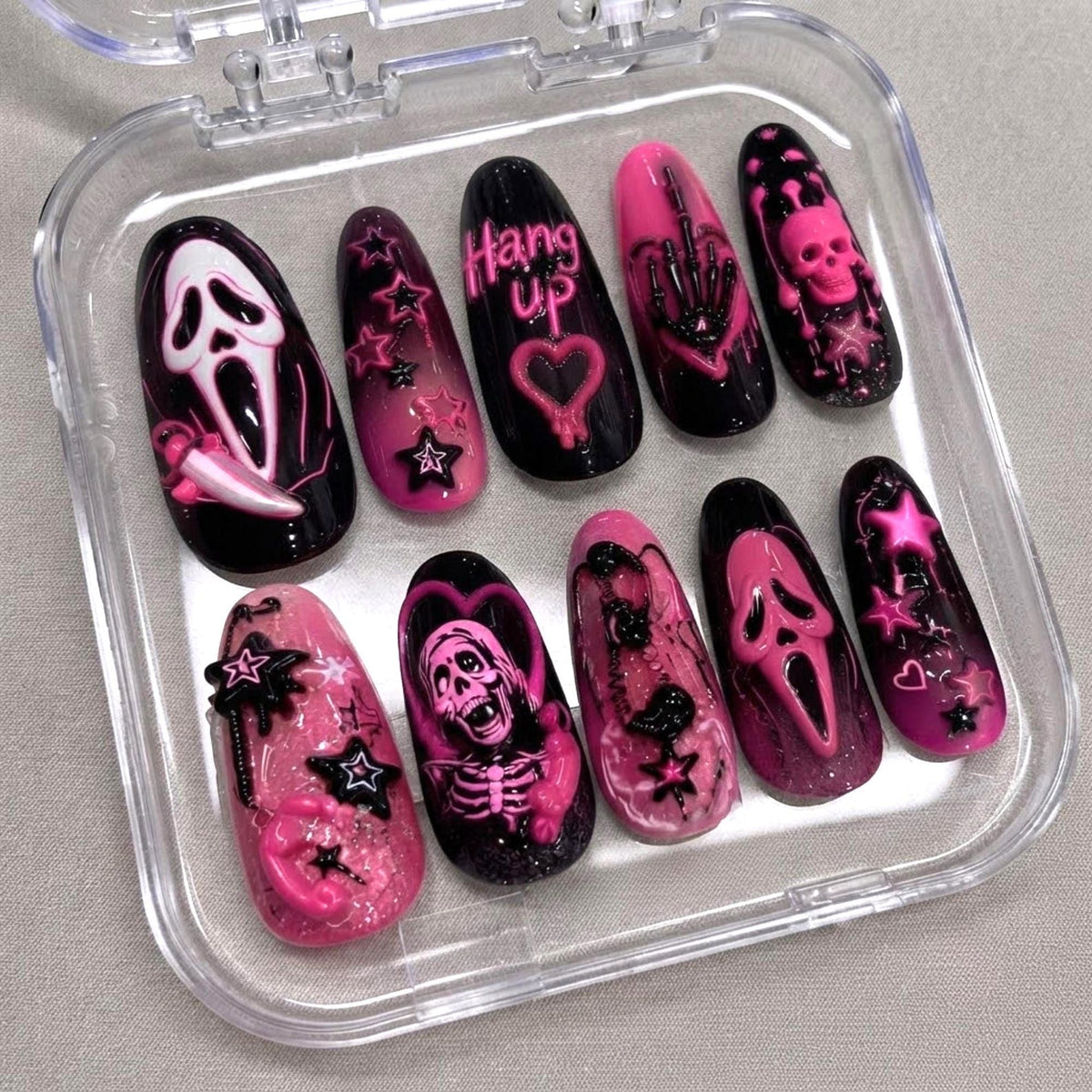 A set of ten press-on nails in a clear plastic case, featuring a black and pink color scheme. Designs include a ghost mask, a skeleton, stars, hearts, and the text "Hang up" with a telephone icon, all rendered in 3D relief.