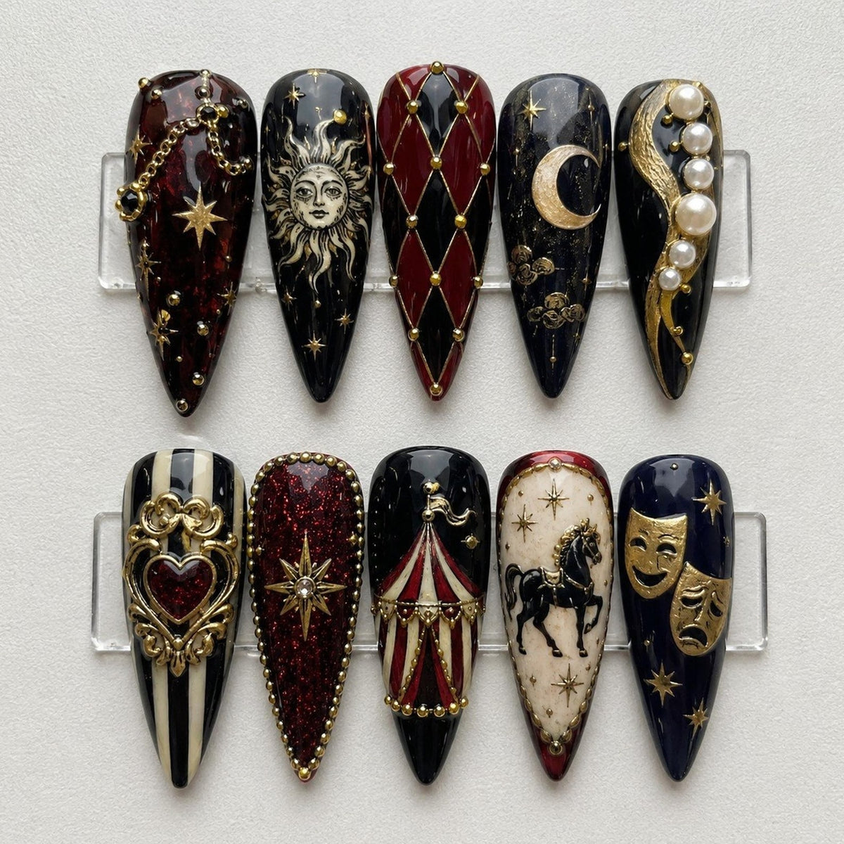 Ten almond-shaped press-on nails featuring intricate designs including a sun face, a crescent moon, a harlequin diamond pattern, a carousel tent, a horse, theater masks, a heart, and gold accents with pearls on black and red backgrounds.