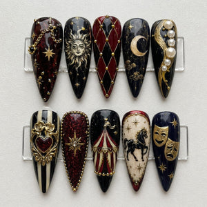 Ten almond-shaped press-on nails featuring intricate designs including a sun face, a crescent moon, a harlequin diamond pattern, a carousel tent, a horse, theater masks, a heart, and gold accents with pearls on black and red backgrounds.