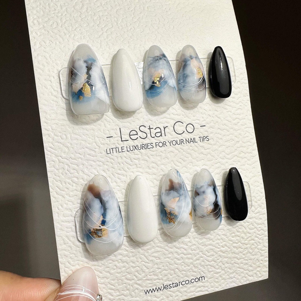 A set of ten almond-shaped press-on nails on a display card labeled LeStar Co. The nails feature a mix of solid white, solid black, and translucent designs with blue and brown marble-like swirls, gold
