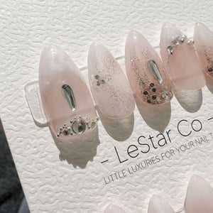 A set of almond-shaped press-on nails in a translucent pale pink hue. The nails feature silver glitter accents, clear teardrop-shaped rhinestones, and small circular crystals. The display card include