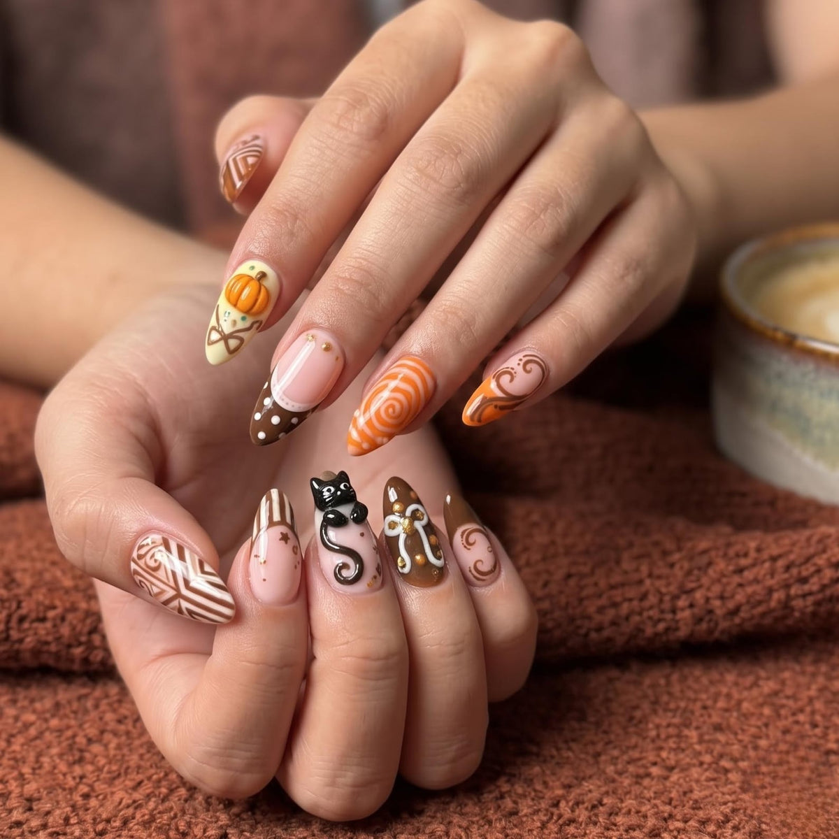 Handmade autumn press on nails with black cat, orange pumpkin, fall leaf and swirl designs in blush pink, brown, black and orange almond shapes.