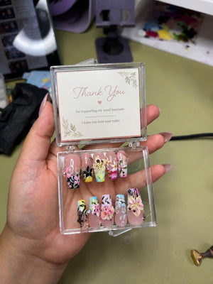 A clear plastic box containing ten press-on nails with floral designs in pink, yellow, and black. The lid features a card with the text Thank You for supporting my small business I hope you love your nails and botanical illustrations.
